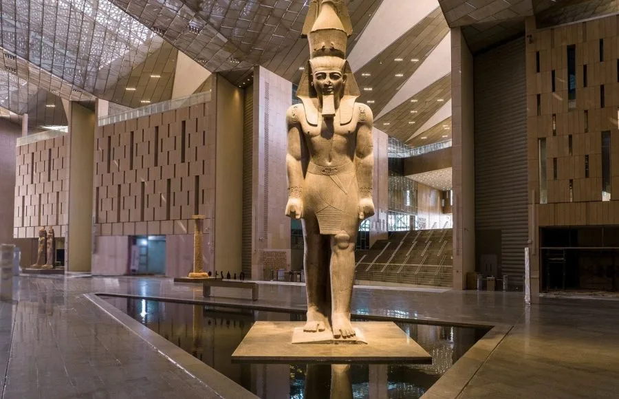 A colossal statue of Ramesses II stands on a podium in the modern, expansive atrium of the Inside The Grand Egyptian Museum GEM, surrounded by sleek architecture and reflective floors.