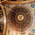 An ornate, intricately decorated dome ceiling of the Mosque of Muhammad Ali Pasha, featuring geometric patterns, gilded accents, and Arabic calligraphy.