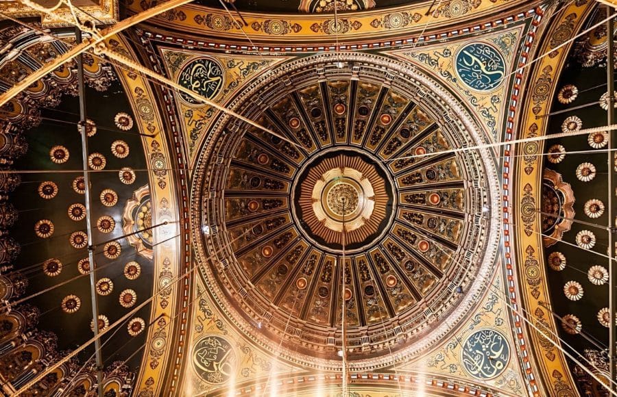 An ornate, intricately decorated dome ceiling of the Mosque of Muhammad Ali Pasha, featuring geometric patterns, gilded accents, and Arabic calligraphy.