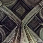An upward-facing close-up of the intricately carved and painted geometric ceiling and arches, and a marble column in the Interior Details at Al Rifai Mosque in Islamic cairo.