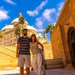 A couple posing happily in a sunny street of Cairo, with the grand domes and minaret of the Muhammad Ali Mosque (Citadel of Saladin) visible in the background.