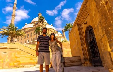 A couple posing happily in a sunny street of Cairo, with the grand domes and minaret of the Muhammad Ali Mosque (Citadel of Saladin) visible in the background.