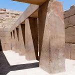 An interior view of the Khafre Valley Temple, Giza, showing a hallway lined with immense, polished, reddish-brown granite pillars leading to the inner chambers under a bright blue sky.