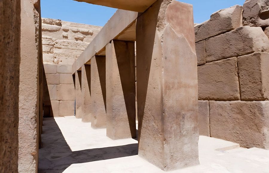 An interior view of the Khafre Valley Temple, Giza, showing a hallway lined with immense, polished, reddish-brown granite pillars leading to the inner chambers under a bright blue sky.