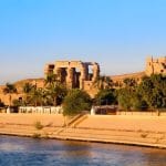 The Kom Ombo Temple at sunset, with its columns and walls as seen from the Nile River, with palm trees and a sandy bank in the foreground.