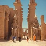 A group of Tourists stands in front of the colossal facade of Luxor Temple, admiring the large statues of Ramses II and the ancient obelisks.