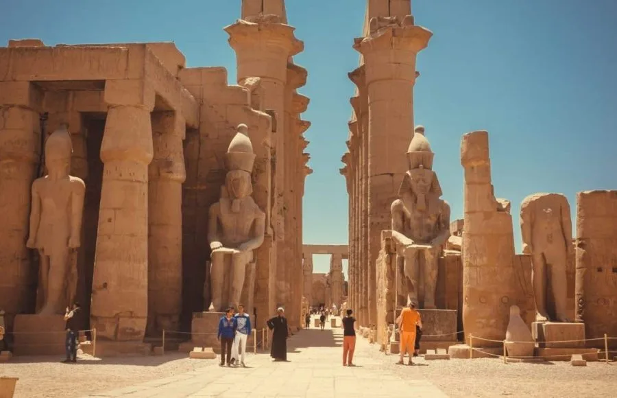 A group of Tourists stands in front of the colossal facade of Luxor Temple, admiring the large statues of Ramses II and the ancient obelisks.