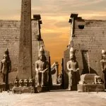 A dramatic Luxor Temple Photo at Night showing the massive entrance pylon, an obelisk, and large statues of Ramses II illuminated under an orange-tinted sky.