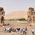 A wide shot of the Luxor Colossi of Memnon, showing the two massive, seated stone statues of Amenhotep III surrounded by a large group of tourists in the desert landscape.