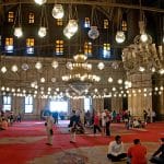 The interior of the Mosque of Muhammad Ali Pasha, with people scattered across a red carpeted floor, illuminated by many hanging spherical glass lamps and large chandeliers.
