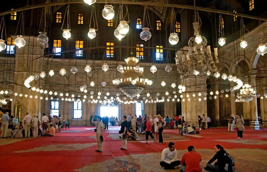 The interior of the Mosque of Muhammad Ali Pasha, with people scattered across a red carpeted floor, illuminated by many hanging spherical glass lamps and large chandeliers.