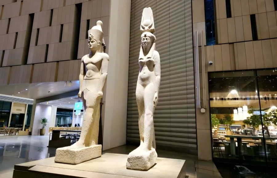 Two ancient Egyptian statues, a male and a female figure, stand impressively in the modern interior of the Grand Egyptian Museum (GEM)