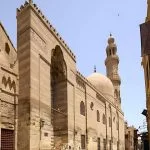 The tall, imposing stone facade of the Mosque Madrasa of Sultan Barquq Complex in Islamic Cairo, featuring a rounded dome and a tall, slender minaret.