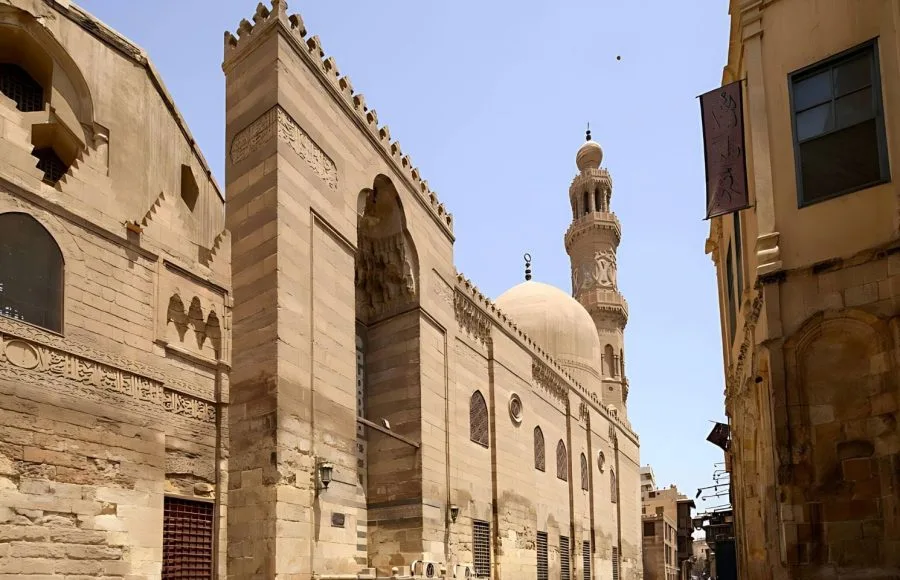 The tall, imposing stone facade of the Mosque Madrasa of Sultan Barquq Complex in Islamic Cairo, featuring a rounded dome and a tall, slender minaret.