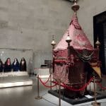 A richly decorated red fabric palanquin or ceremonial canopy on display in a museum hall, with several dark robed female figures in glass cases behind it.