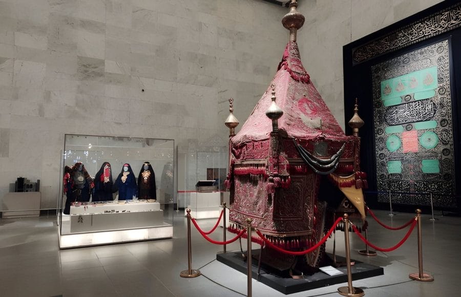 A richly decorated red fabric palanquin or ceremonial canopy on display in a museum hall, with several dark robed female figures in glass cases behind it.