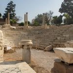 The sunlit, tiered seating and stone ruins of The Roman Amphitheatre next to a column capital at the archaeological site of Pompeys Pillar in Alexandria.