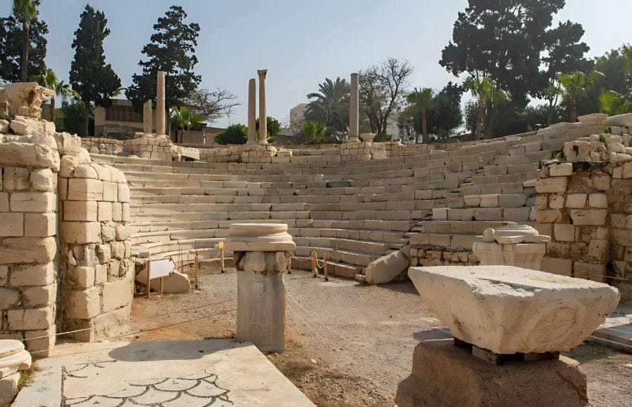 The sunlit, tiered seating and stone ruins of The Roman Amphitheatre next to a column capital at the archaeological site of Pompeys Pillar in Alexandria.