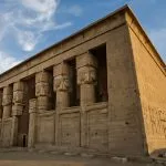 A wide shot of the majestic Ptolemaic Temple of Hathor at Dendera, featuring large columns with Hathoric capitals under a bright sky.