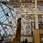 The colossal statue of Ramses II stands in the Grand Egyptian Museum, against a towering modern glass and steel structure that forms the museum's interior facade.