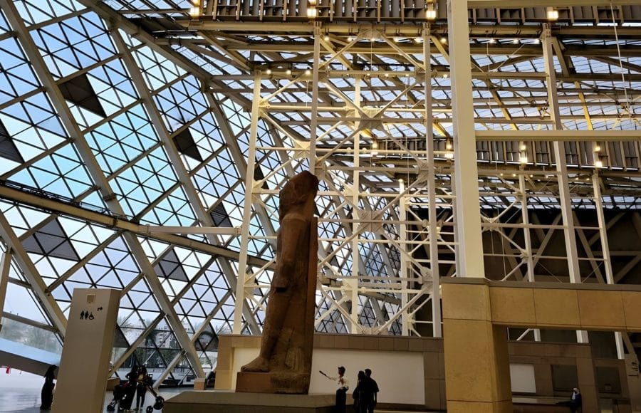 The colossal statue of Ramses II stands in the Grand Egyptian Museum, against a towering modern glass and steel structure that forms the museum's interior facade.
