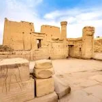 A wide-angle shot of the partially ruined Edfu Temple, showing its imposing walls and the ruins of surrounding structures under a bright sky.