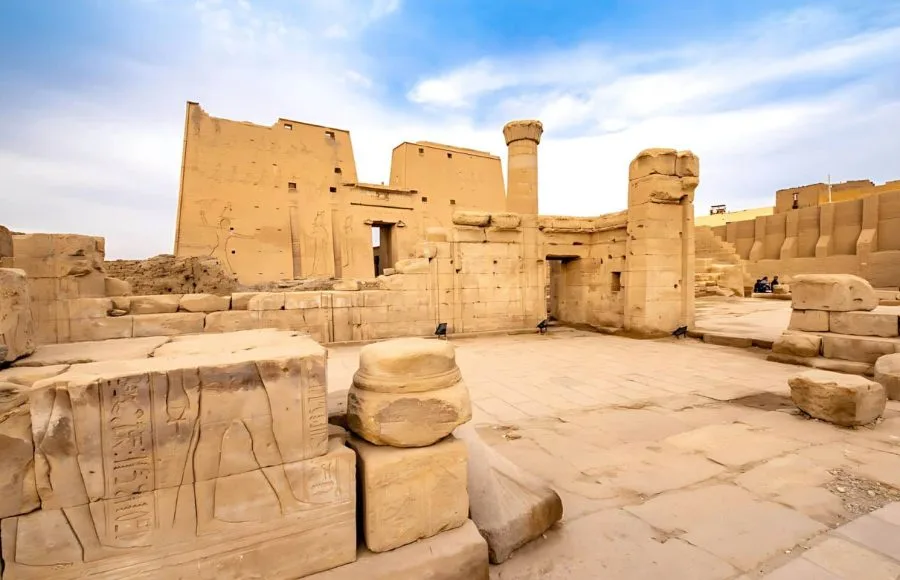 A wide-angle shot of the partially ruined Edfu Temple, showing its imposing walls and the ruins of surrounding structures under a bright sky.