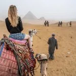A Solo Female Traveler with long blonde hair, sitting on a camel and looking toward the Pyramids of Giza in the background, led by a guide in the desert sand.