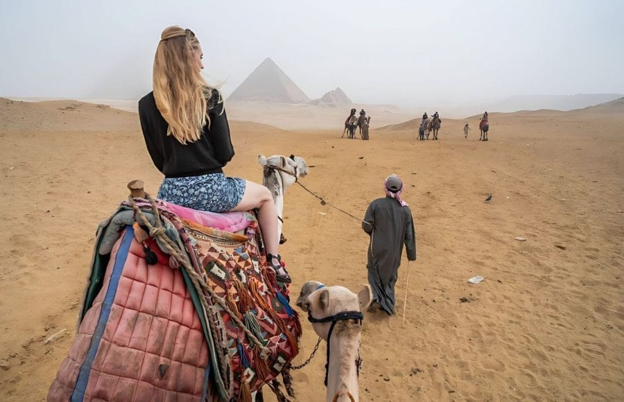 A Solo Female Traveler with long blonde hair, sitting on a camel and looking toward the Pyramids of Giza in the background, led by a guide in the desert sand.