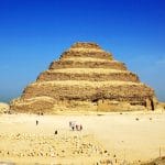 The ancient stepped structure of the Step Pyramid Saqqara Giza Tour rising against a clear blue sky in the desert, with scattered ruins and visitors at its base.