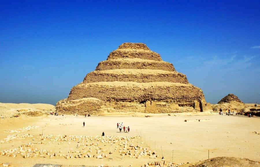 The ancient stepped structure of the Step Pyramid Saqqara Giza Tour rising against a clear blue sky in the desert, with scattered ruins and visitors at its base.