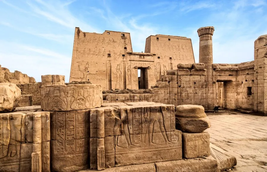 A stone building with carvings on it with Temple of Edfu in the background