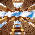 An upward view of the colossal, carved columns inside the Hypostyle Hall of the Temple of Karnak Columns Luxor, showing intricate reliefs against a bright blue sky.