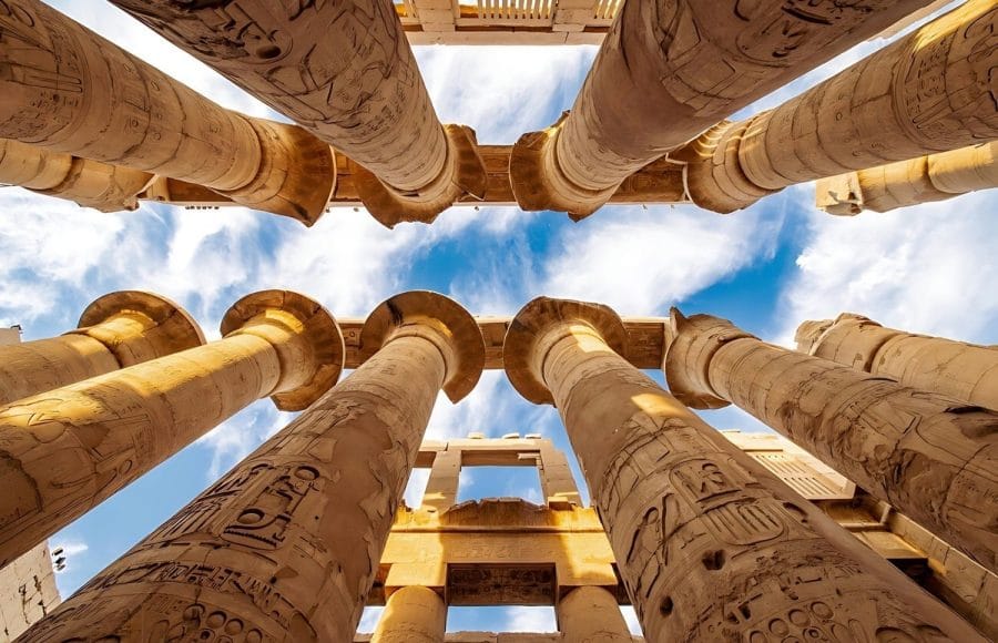 An upward view of the colossal, carved columns inside the Hypostyle Hall of the Temple of Karnak Columns Luxor, showing intricate reliefs against a bright blue sky.