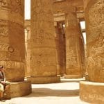 A tourist sits at the base of one of the colossal, carved Temple of Karnak Columns in Luxor, Egypt, gazing up at the ancient Hieroglyphics.
