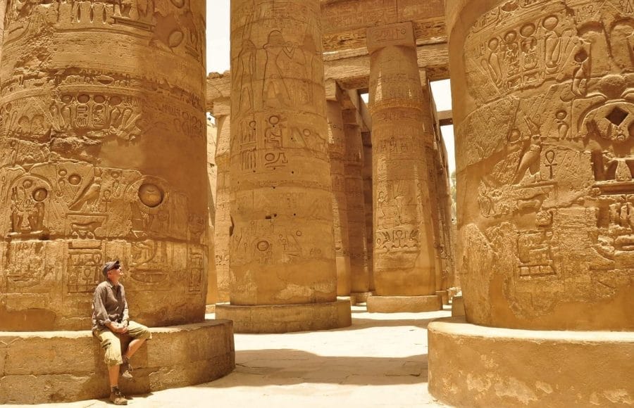 A tourist sits at the base of one of the colossal, carved Temple of Karnak Columns in Luxor, Egypt, gazing up at the ancient Hieroglyphics.