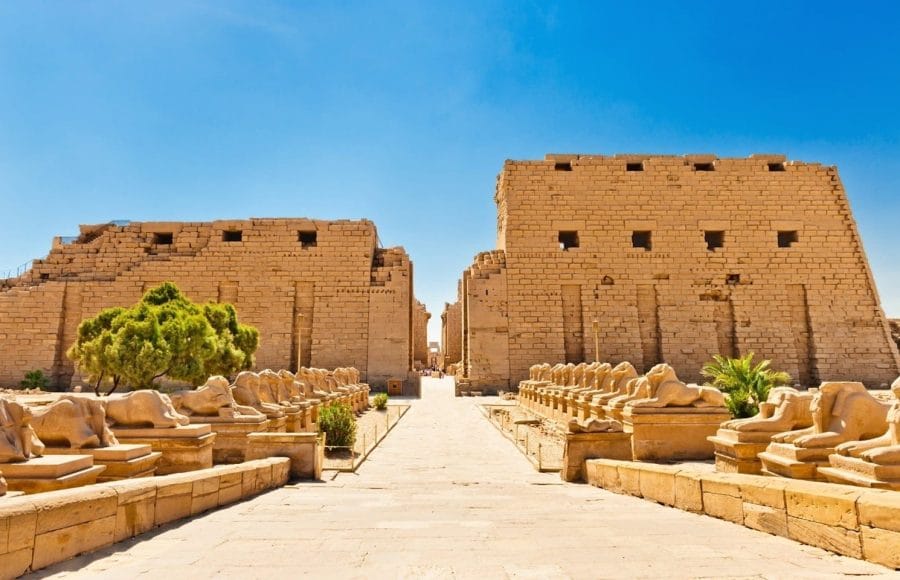 The grand entrance to the Temple of Luxor, showing the processional pathway lined with rows of sphinx statues and the massive stone pylon towers in the background.