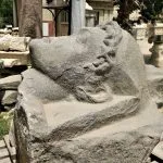 A large, weathered, carved stone fragment of a Colossal head of a Ptolemy or the Grey granite head of Mark Antony, displayed outdoors in the courtyard of the Greco-Roman Museum.