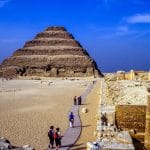The ancient Step Pyramid of Djoser at Saqqara in Cairo, with a wooden walkway leading towards it across a sandy desert landscape.
