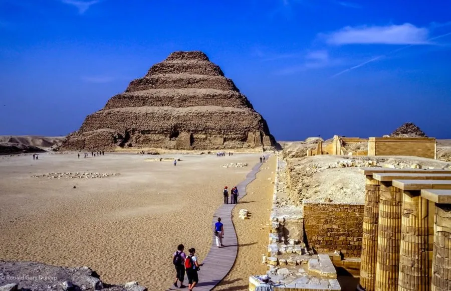 The ancient Step Pyramid of Djoser at Saqqara in Cairo, with a wooden walkway leading towards it across a sandy desert landscape.