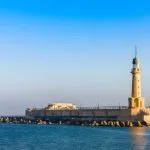 A modern lighthouse structure built on a stone breakwater, known as The Lighthouse at Alexandria, Egypt, standing tall over the calm blue Mediterranean Sea.