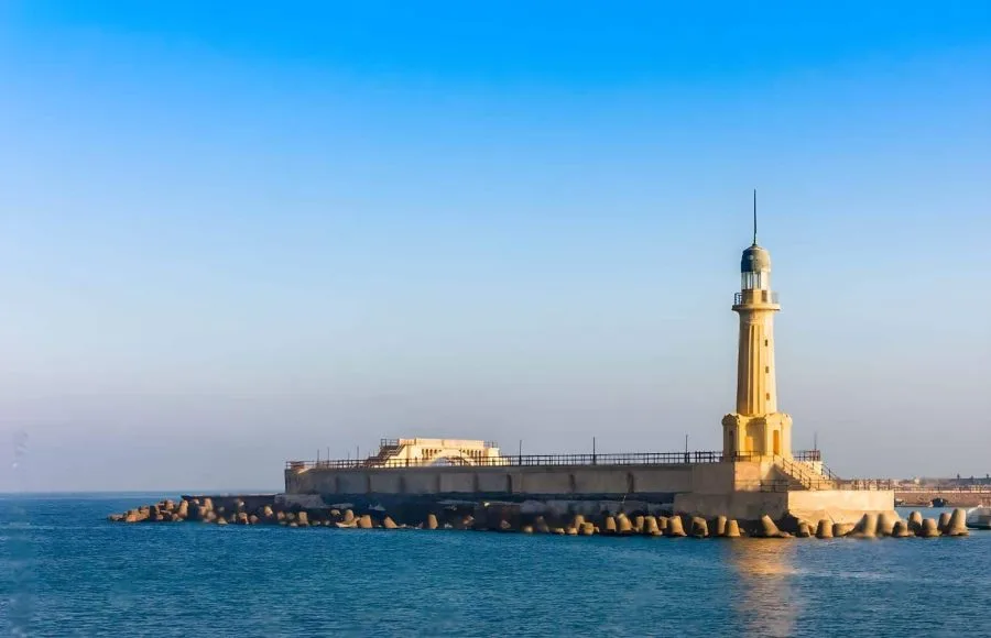 A modern lighthouse structure built on a stone breakwater, known as The Lighthouse at Alexandria, Egypt, standing tall over the calm blue Mediterranean Sea.