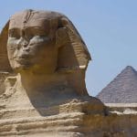 A close up view of the Great Sphinx of Giza, showing its detailed facial features and body, with one of the pyramids visible in the background.
