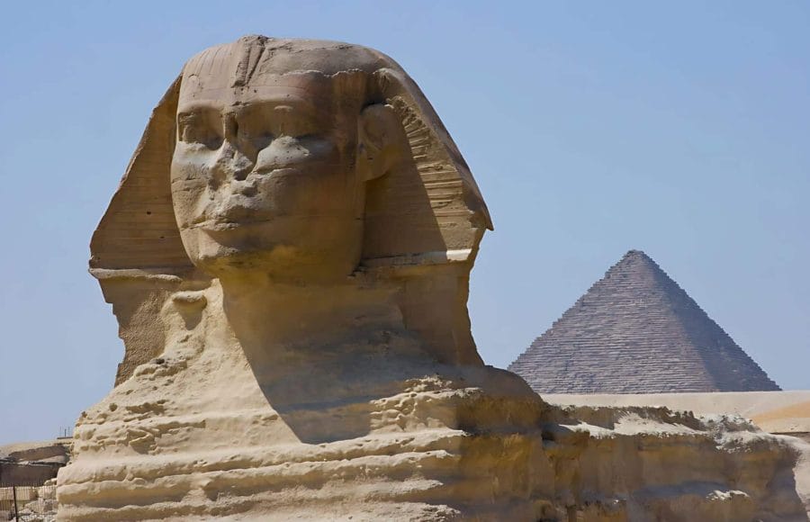A close up view of the Great Sphinx of Giza, showing its detailed facial features and body, with one of the pyramids visible in the background.