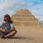 A man wearing a cowboy hat sits on the desert ground, gazing up at the massive, stepped structure of The Step Pyramid of Djoser against a cloudy blue sky.