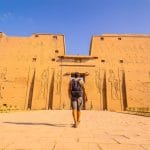 A tourist walks towards the grand entrance of the Temple of Edfu, a well-preserved ancient temple with carved stone facades.