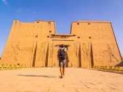 A tourist walks towards the grand entrance of the Temple of Edfu, a well-preserved ancient temple with carved stone facades.