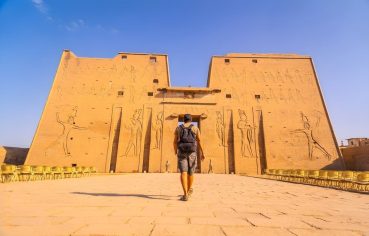 A tourist walks towards the grand entrance of the Temple of Edfu, a well-preserved ancient temple with carved stone facades.
