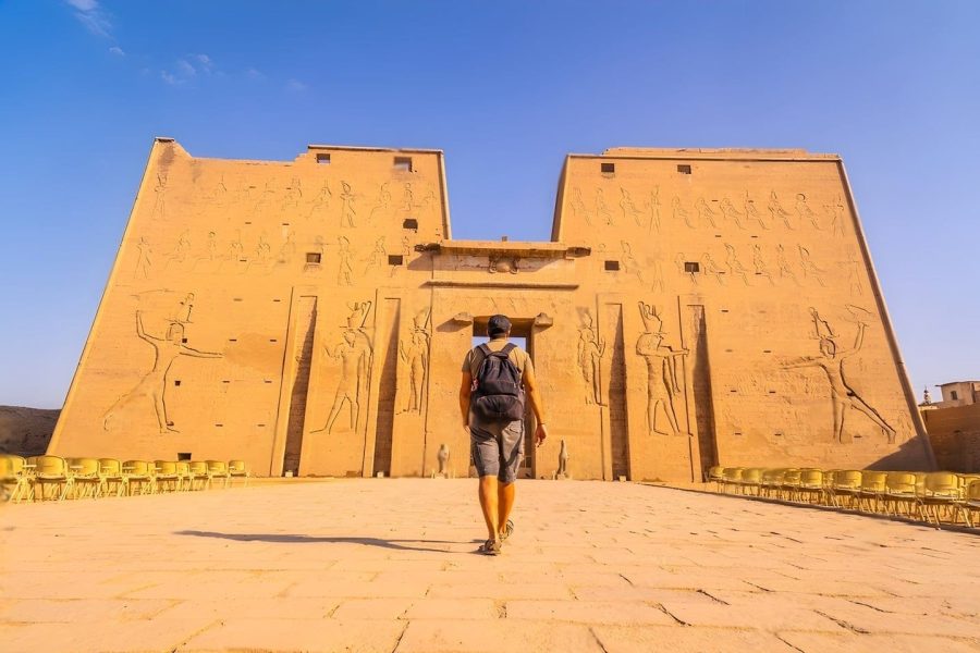 A Tourist Walks Towards The Grand Entrance Of The Temple Of Edfu, A Well-Preserved Ancient Temple With Carved Stone Facades.
