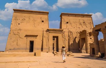 A tourist walking towards the beautifully decorated pylons of the Philae Temple under a bright sky with scattered clouds.